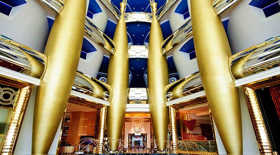 Grand atrium of Burj Al Arab with towering gold columns, blue ceiling, and ornate golden-framed doorways leading to elegant i