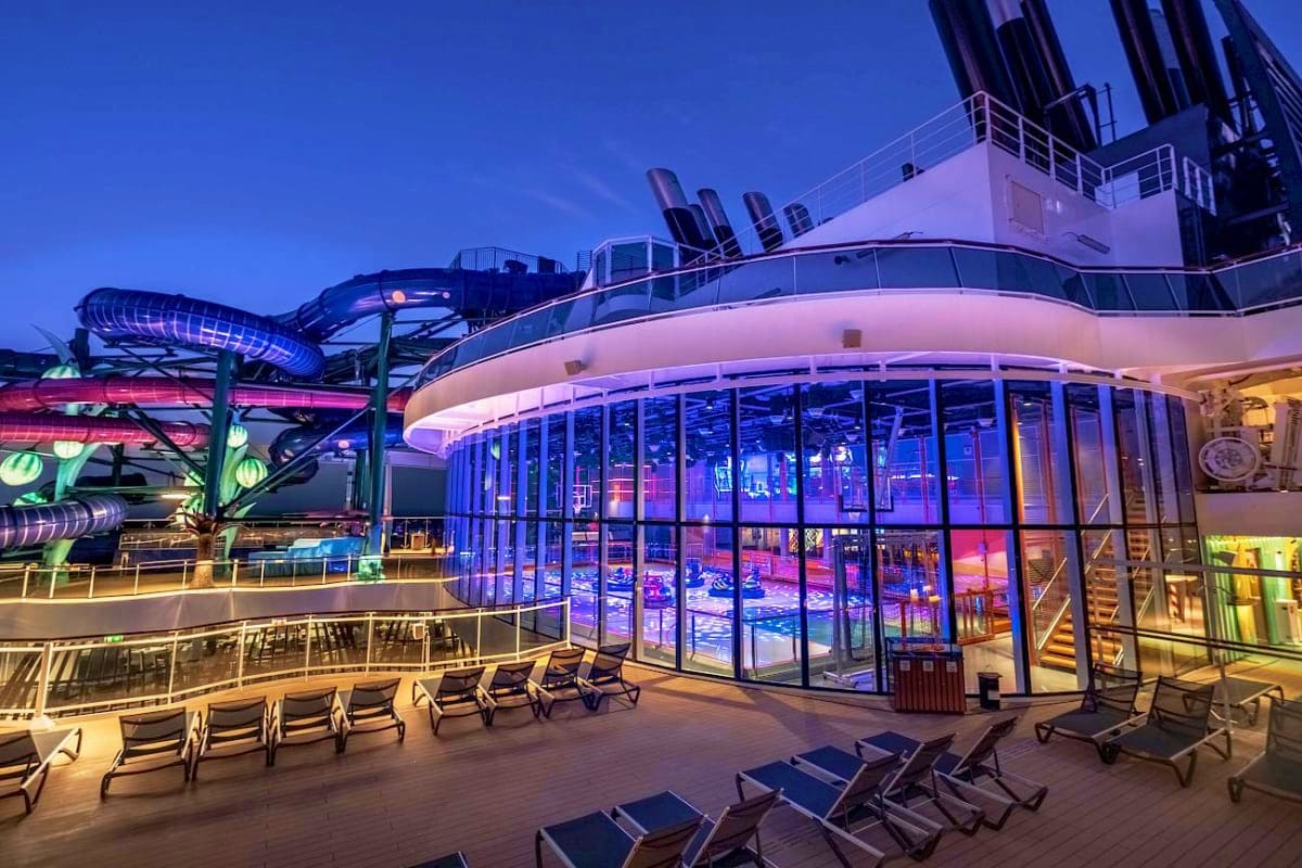 MSC World Europa Barcelona Roundtrip Cruise Illuminated cruise ship deck at dusk with lounge chairs, water slide, and blue-lit indoor pool visible through large windows