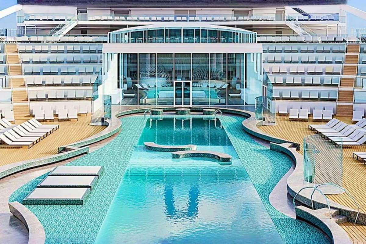 Modern cruise ship deck with Olympic-sized swimming pool, lounge chairs, and multi-story white building with floor-to-ceiling