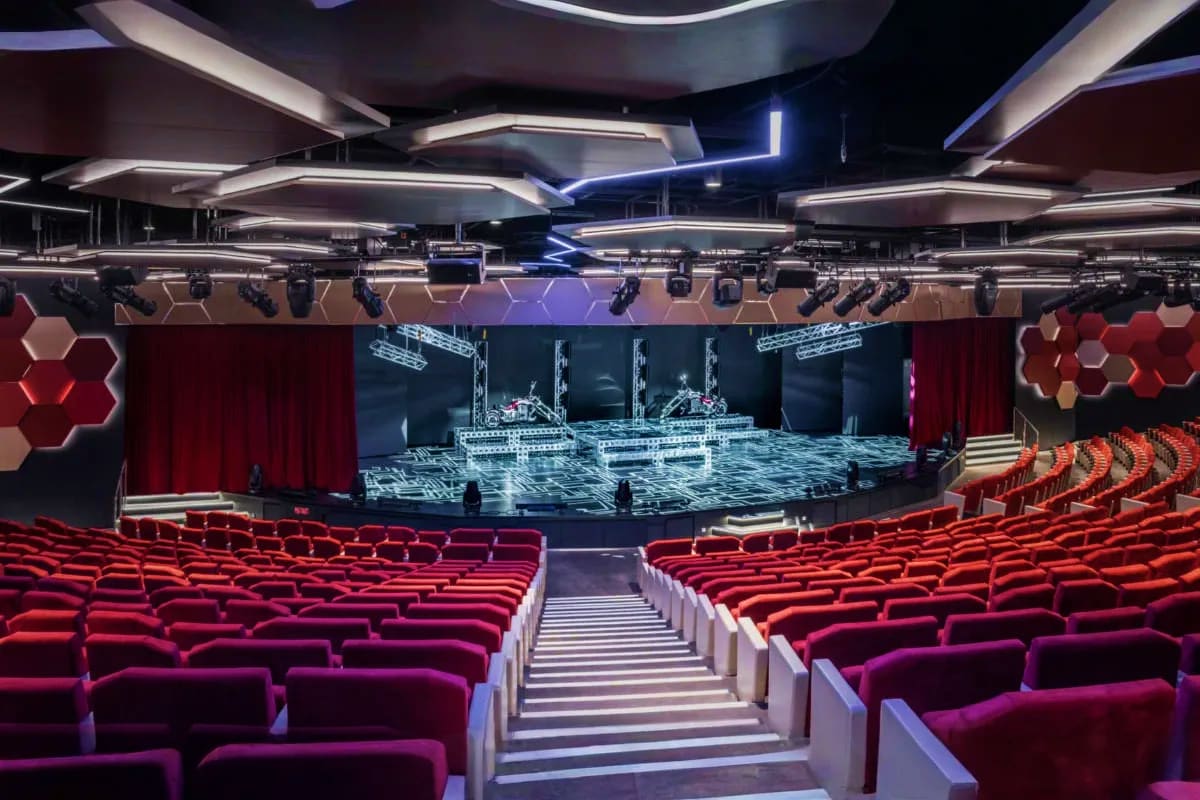 Modern cruise ship theater with red velvet seats, illuminated stage, hexagonal ceiling panels, and professional lighting rig