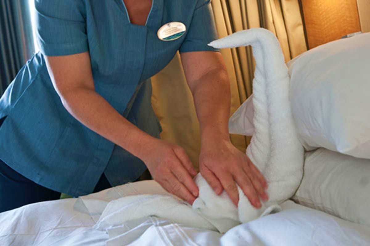 Cruise staff member in blue uniform smoothing white bedding in a cabin with gold curtains in background