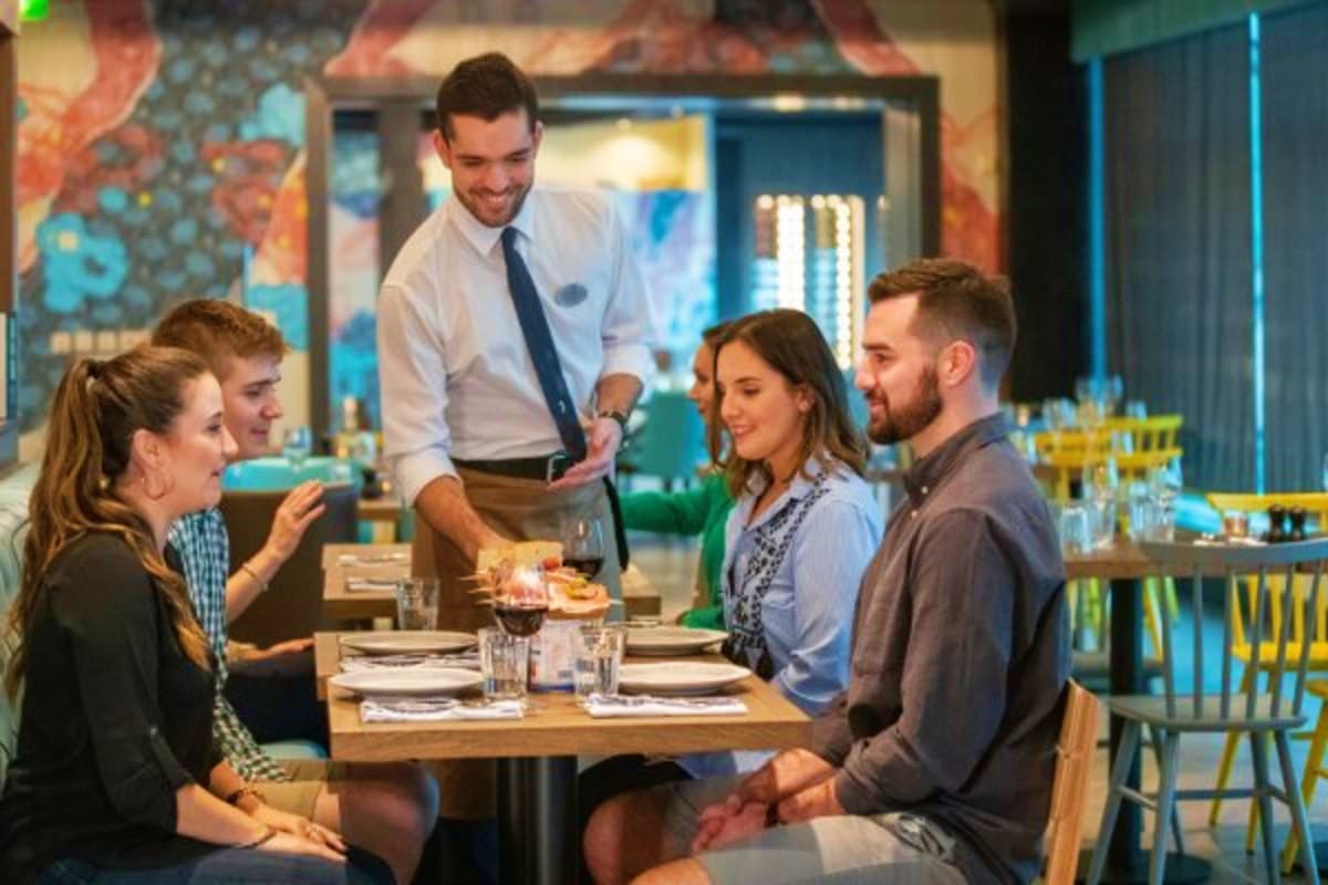 Server taking order from four diners at wooden table in modern restaurant with colorful wall art and blue lighting
