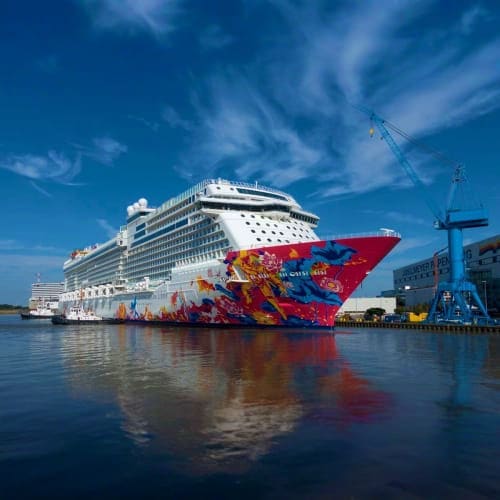 Large colorful cruise ship with red hull docked at industrial port under blue sky with white clouds