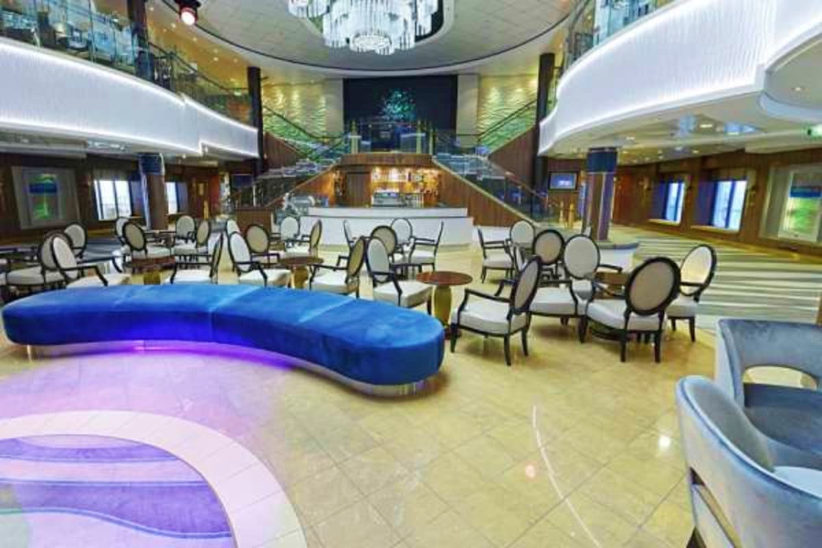 Modern cruise ship atrium with blue curved seating, elegant dining chairs, polished floors, and multi-level balconies with co