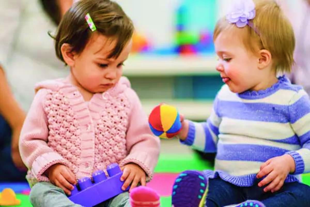 Two toddlers playing with colorful toys and building blocks on a playroom floor in a daycare or preschool setting.