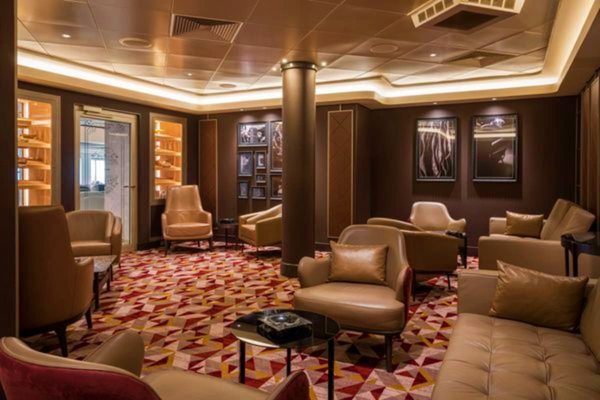 Luxury cruise ship lounge with beige leather seating, geometric patterned carpet, dark wood paneling, and ambient ceiling lig