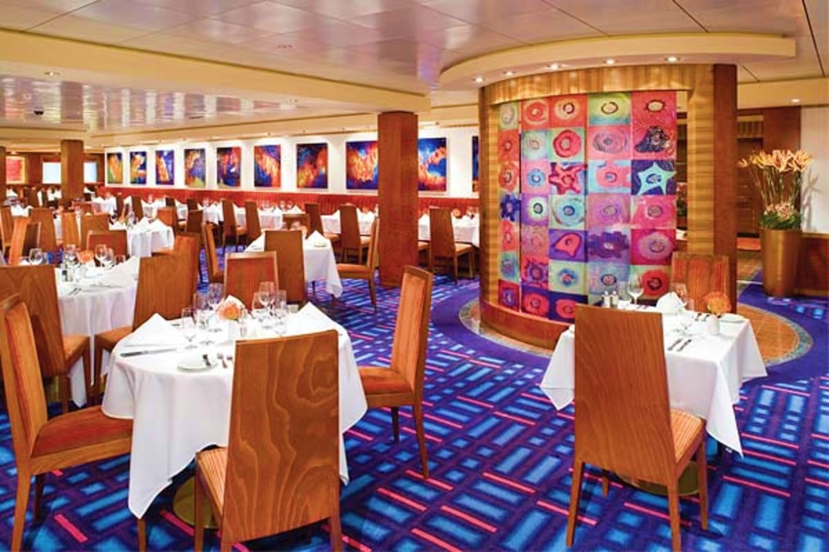Elegant cruise ship dining room with round tables set with white linens, wood chairs, blue patterned carpet, and colorful Asi