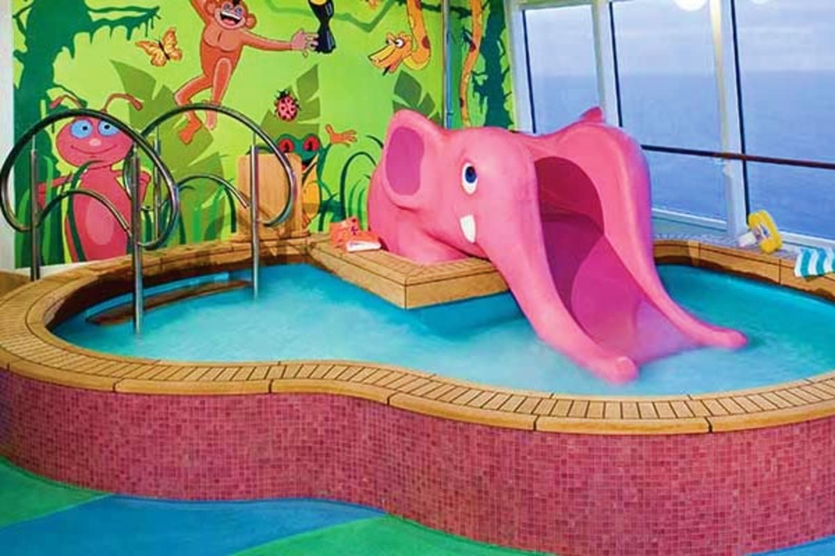 Pink elephant-shaped water slide in children's pool area with colorful jungle mural and ocean view windows on cruise ship