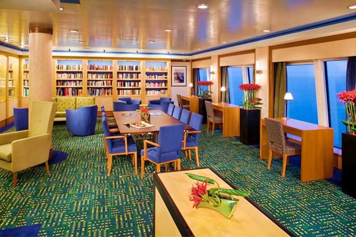 Modern cruise ship library lounge with bookshelves, dining table, comfortable seating, ocean views through large windows, and