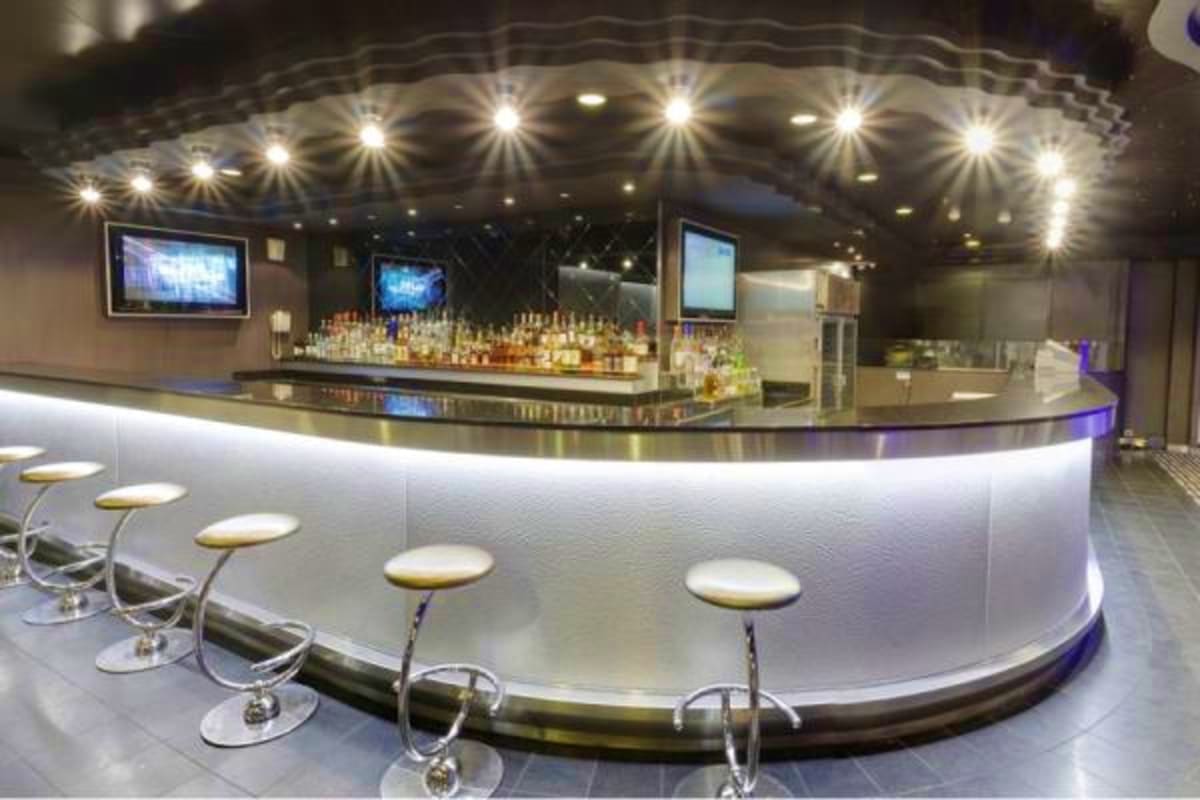 Modern cruise ship bar with curved white counter, bar stools, illuminated shelving with liquor bottles, TV screens, and decor