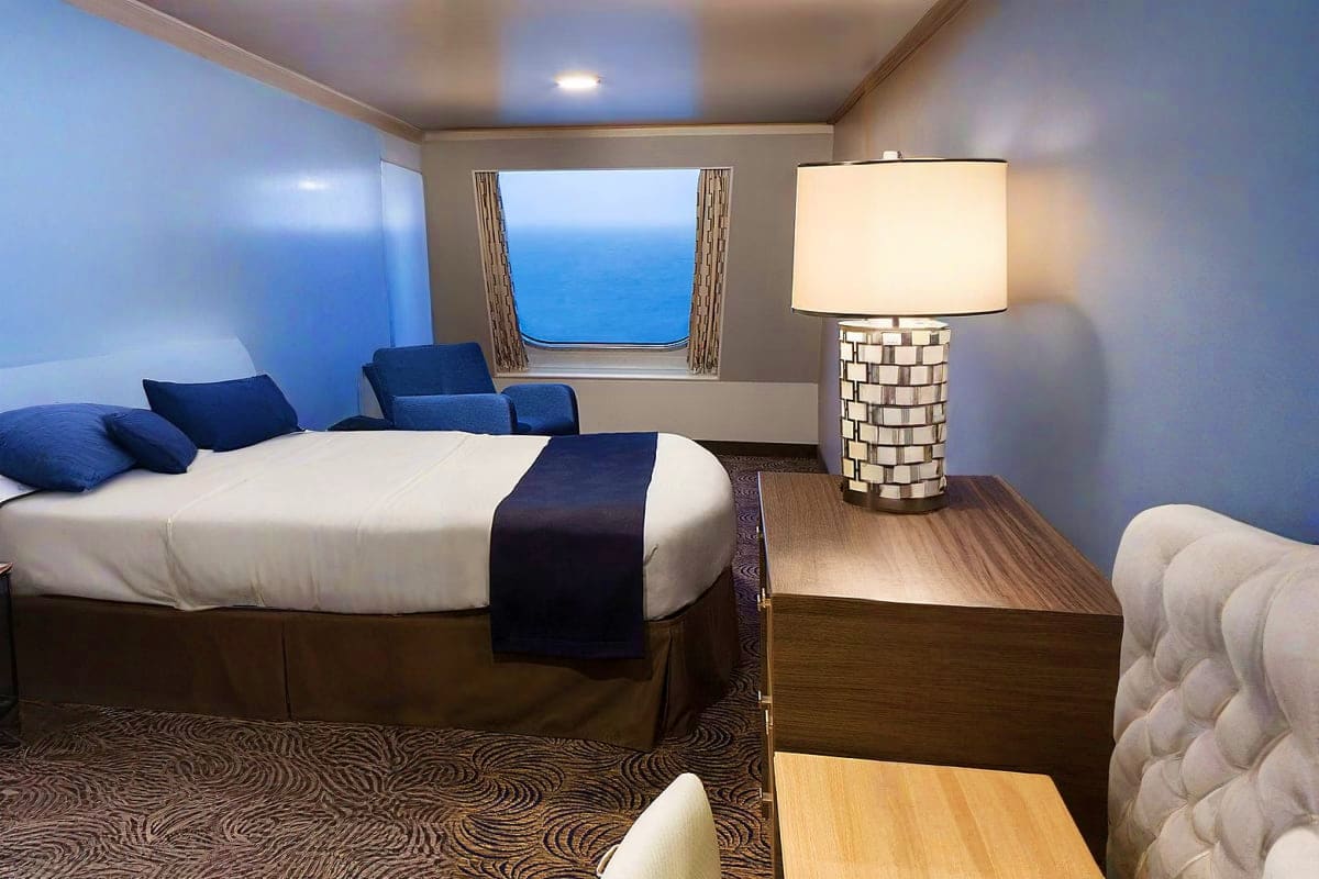 Odyssey of the Seas Bahamas Signature Cruise ship stateroom with ocean view window, queen bed, blue armchair, table lamp, and modern furnishings overlooking blue s