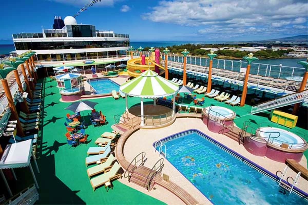 Aerial view of Norwegian Jade cruise ship pool deck with swimming pools, water slides, lounge chairs, and coastal scenery in 