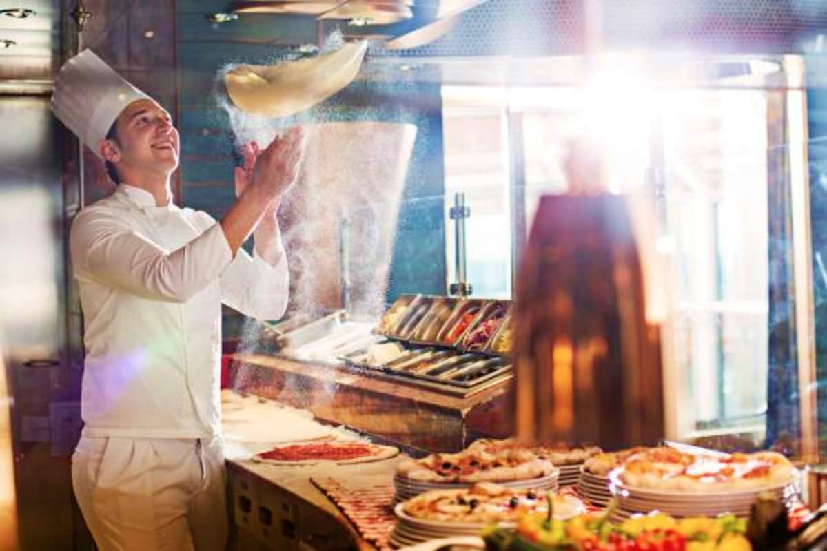 Chef tossing pizza dough in galley kitchen with finished pizzas and fresh ingredients displayed on counter