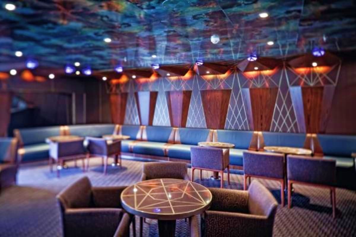 Modern cruise ship lounge with blue seating, geometric patterned ceiling, ambient lighting, and decorative table with geometr