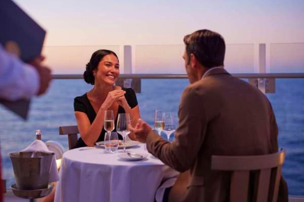 Couple dining at a candlelit table on a ship deck overlooking the ocean at sunset during a cruise dinner service
