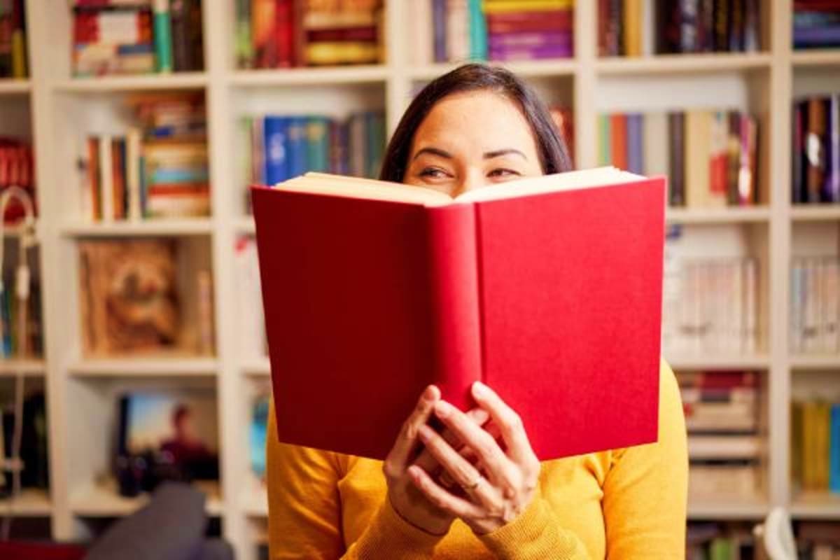 Woman in yellow shirt reading red book in front of white bookshelf in library