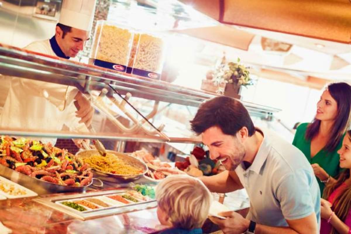Family ordering food at casual buffet restaurant counter with staff member serving pizza and various dishes