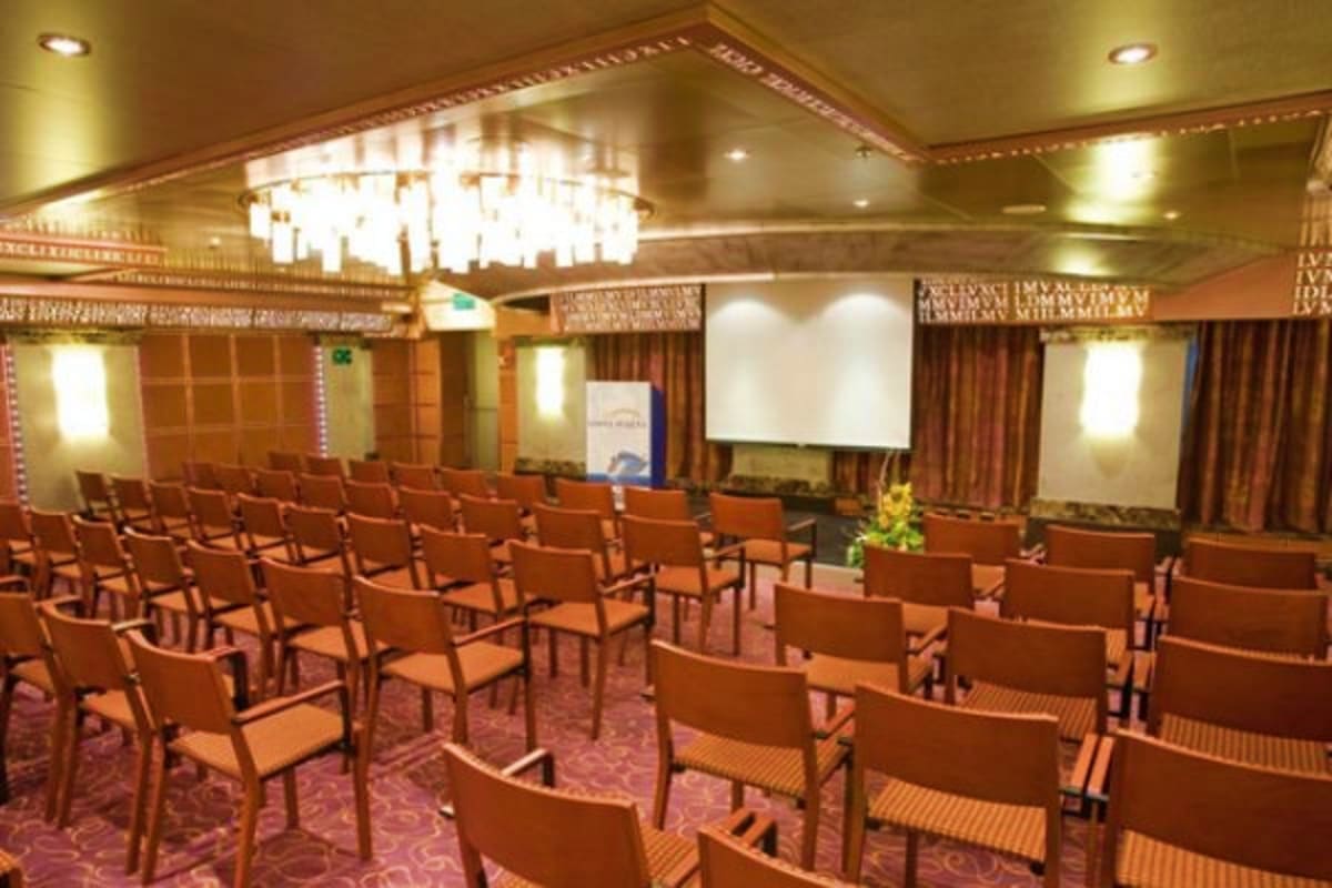 Elegant auditorium on Costa Fascinosa cruise ship with rows of orange chairs, crystal chandelier, projection screen, and warm