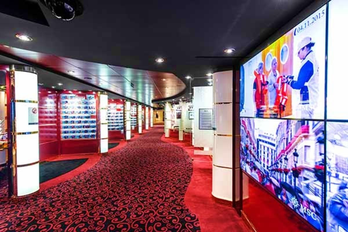 Modern cruise ship corridor with red patterned carpet, illuminated display cases, and large digital screens showing colorful 