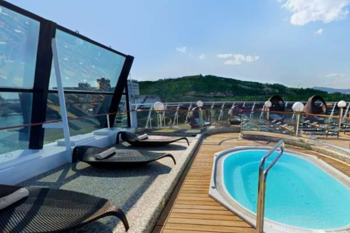 Cruise ship pool deck with blue swimming pool, lounge chairs, and river landscape with green hills in background