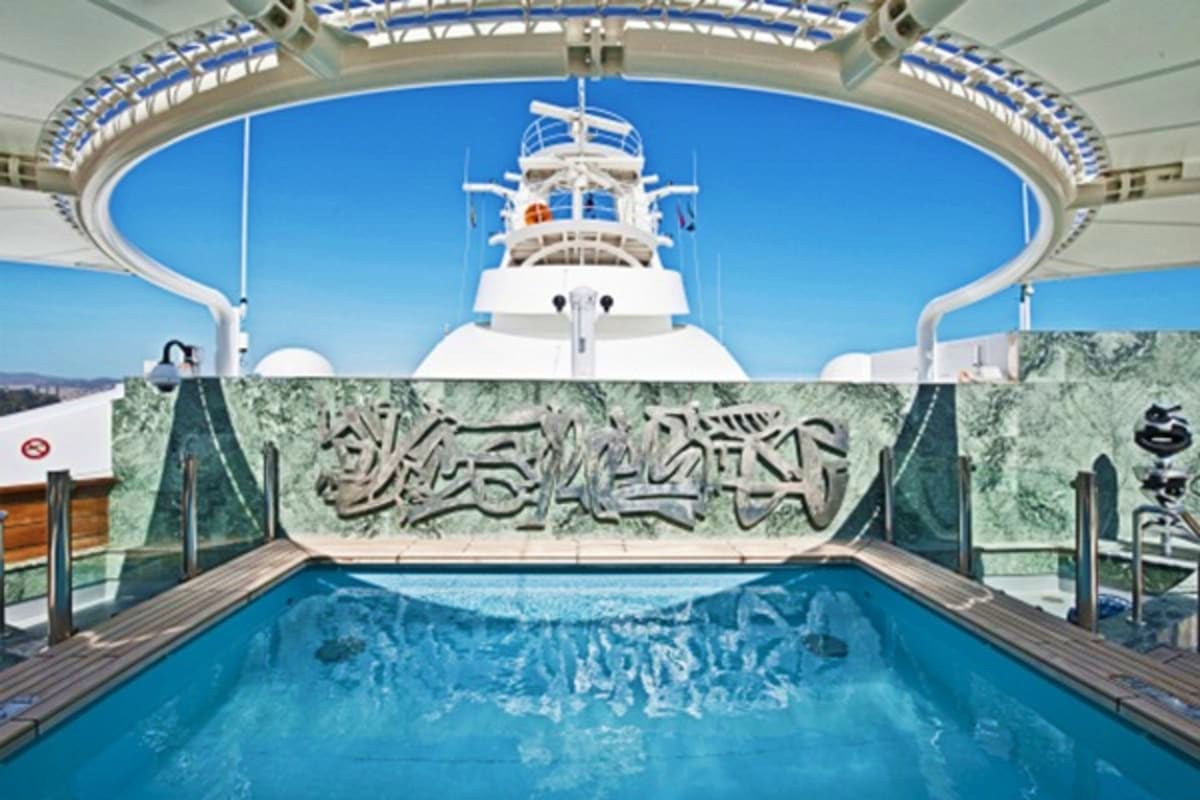 Luxury cruise ship pool deck with blue water, white vessel visible above, decorative stone relief wall, and ocean vista under