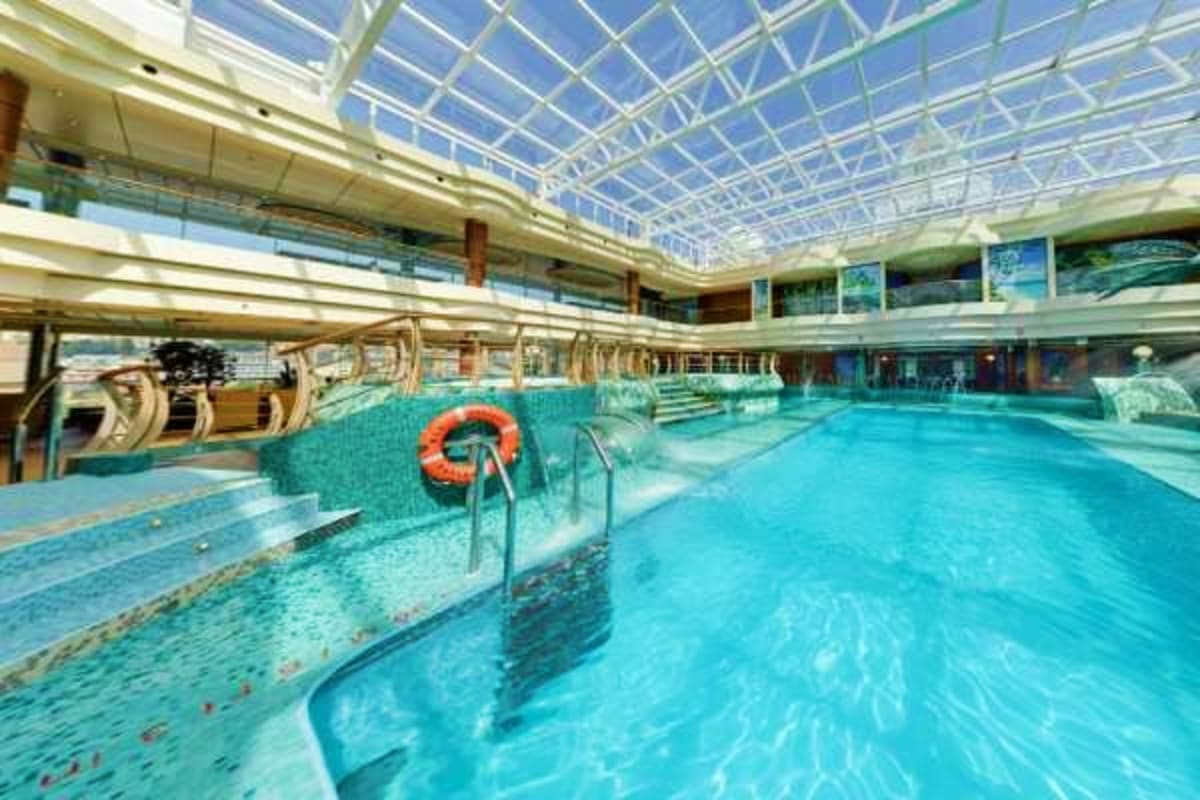 Indoor swimming pool with blue water, orange life ring, metal railings, and glass ceiling with natural light on cruise ship