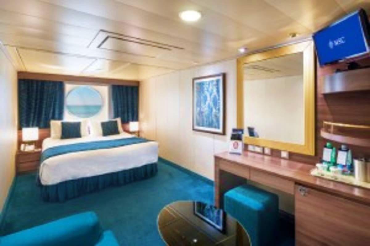 Luxurious cruise ship stateroom with queen bed, sitting area, porthole window with ocean view, and modern vanity with mirror 