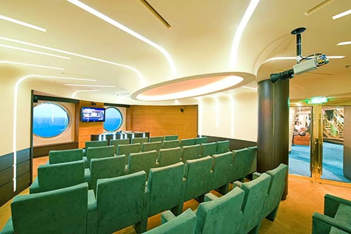Modern cruise ship theater interior with green seats, wood paneling, circular portholes, and illuminated ceiling lighting fix
