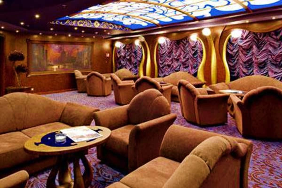 Luxury cruise ship theater seating area with tan upholstered armchairs, decorative patterned curtains, illuminated ceiling, a