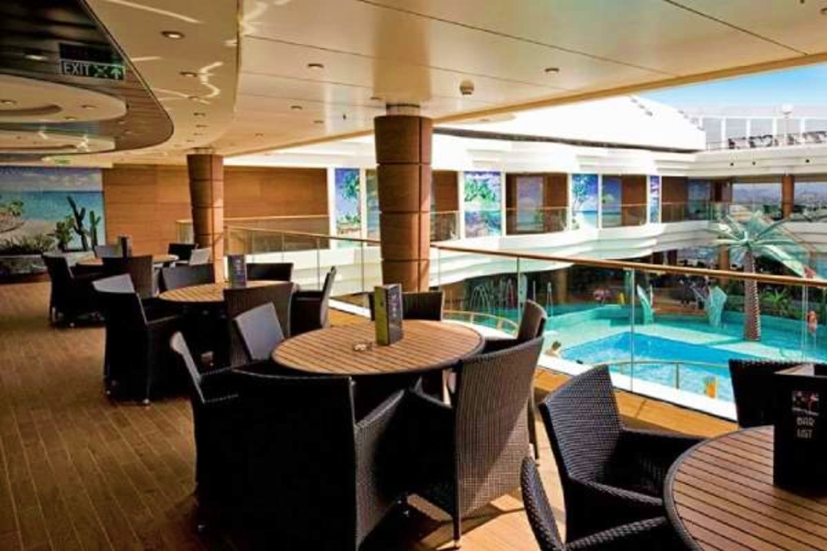 Cruise ship deck lounge area with dining tables, swimming pool, and ocean view through large windows