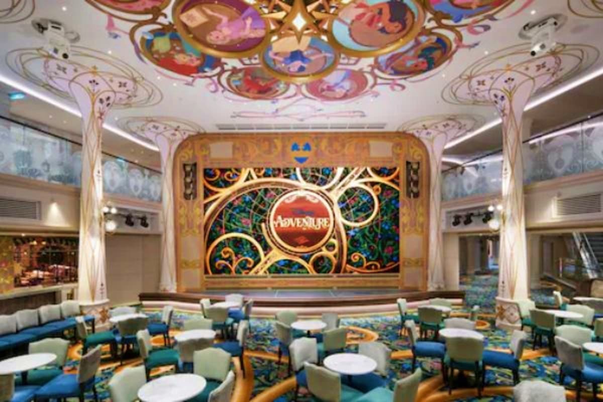 Ornate Disney theater interior with colorful ceiling frescoes, gold-trimmed stage backdrop, and dining tables with blue patte