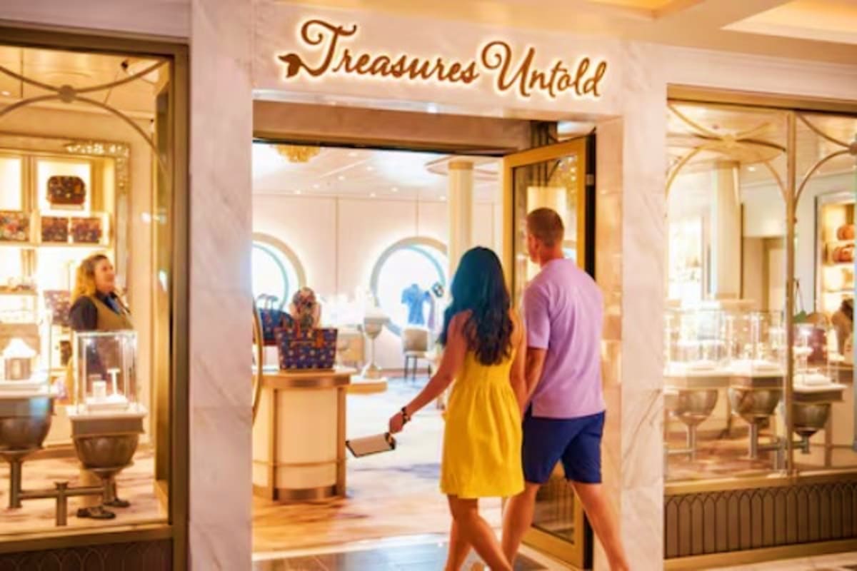 Couple entering Treasures Untold jewelry store on Disney cruise ship with illuminated storefront and ocean portholes visible 