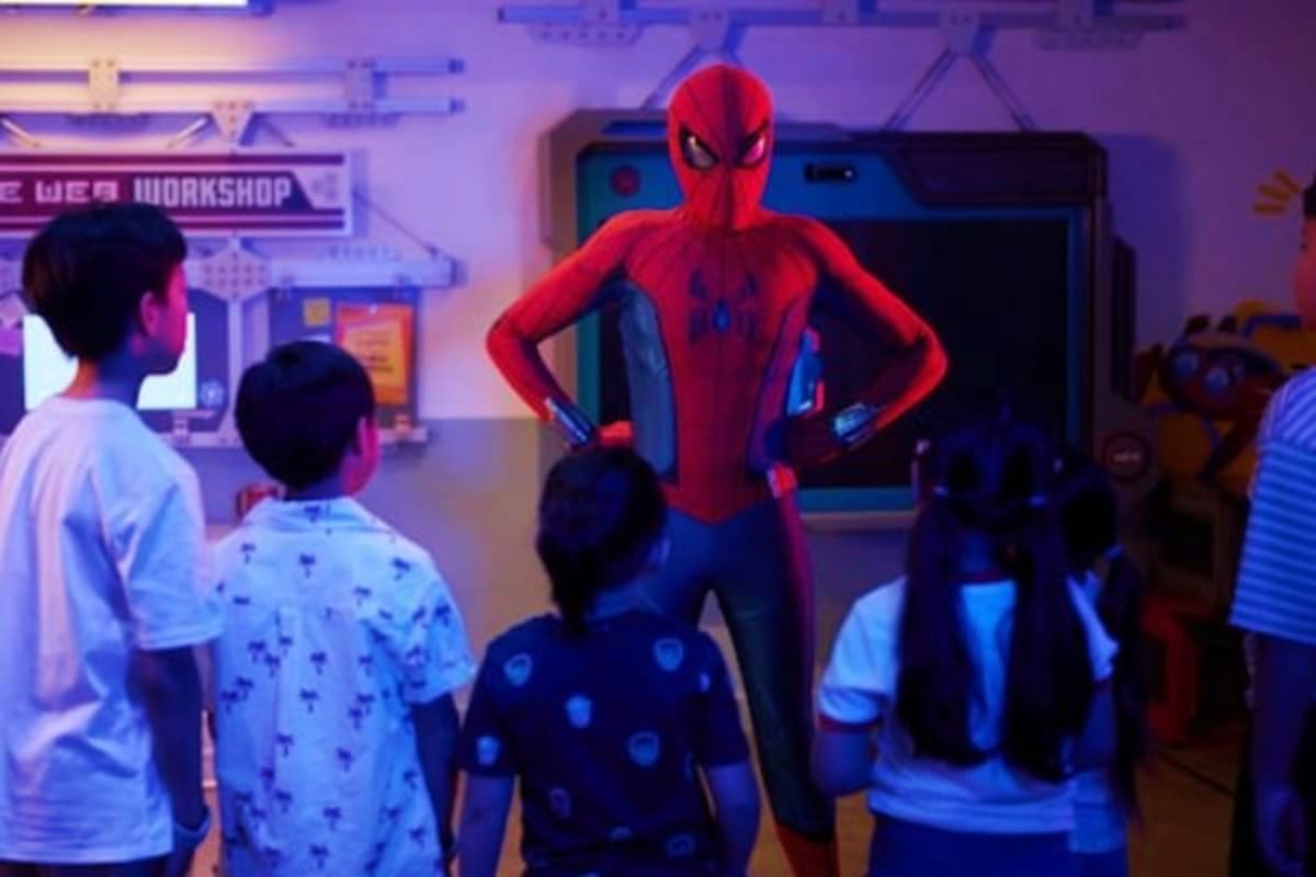 Spider-Man character in red and blue suit standing with hands on hips facing group of children in museum or exhibition space 