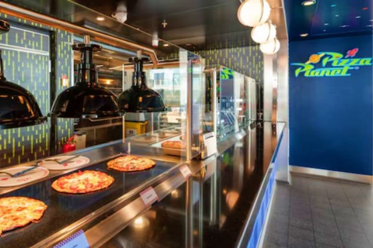 Modern pizza restaurant interior with fresh pizzas under heat lamps and "Pizza Planet" signage on blue wall