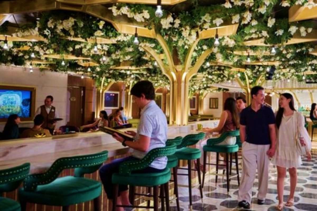 Modern hotel lobby with green velvet bar stools, golden tree-shaped ceiling structures with hanging white flowers, and guests