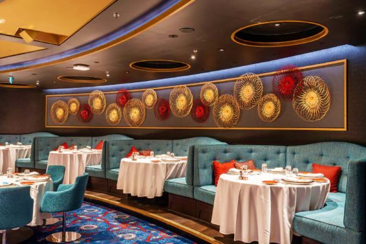 Upscale cruise ship dining room with teal banquette seating, round tables with cream linens, and decorative gold and red wall