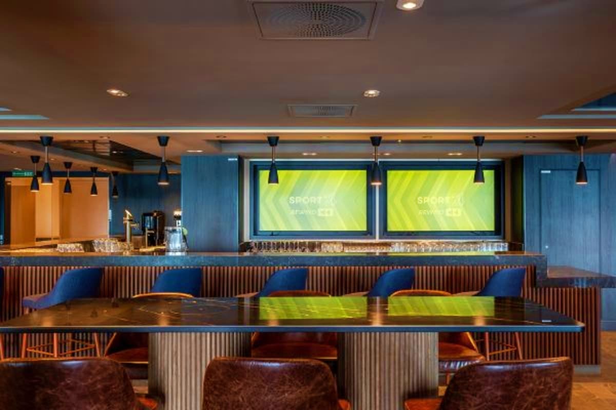 Modern sports bar interior with large screens displaying green content, blue chairs, wooden counter, and pendant lighting