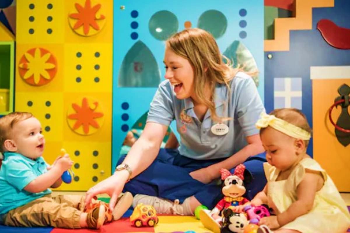 Childcare provider engaged with two toddlers playing with colorful toys in a bright, cheerful classroom with Disney-themed wa