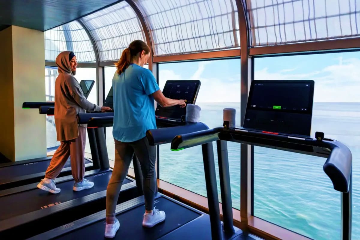 Two women exercising on treadmills in a modern gym with ocean views through large windows overlooking the sea.