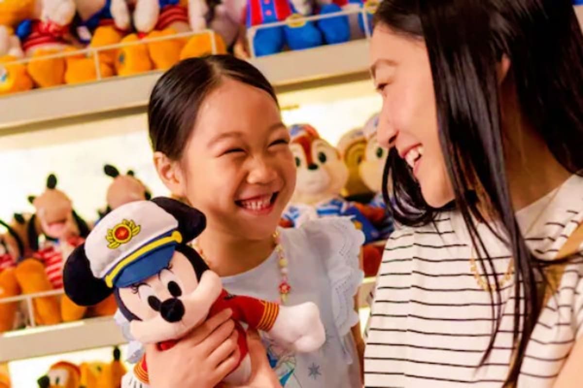 Young girl holding Mickey Mouse plush toy smiling at woman in Disney store with colorful character merchandise displays in ba