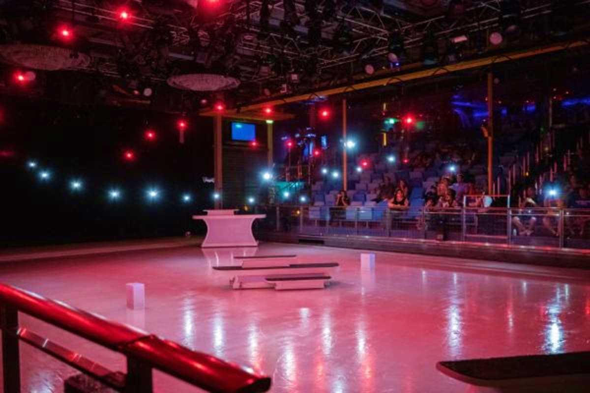 Modern circus arena with pink illuminated floor, red and white stage lighting, spectator seating, and metal railings during e