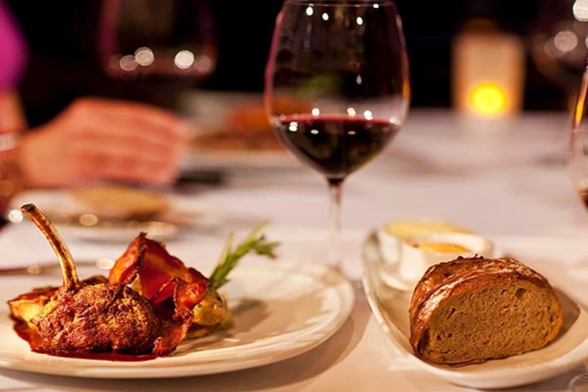 Fine dining table setting with red wine glass, roasted lamb chop with rosemary, and sliced bread on white plates