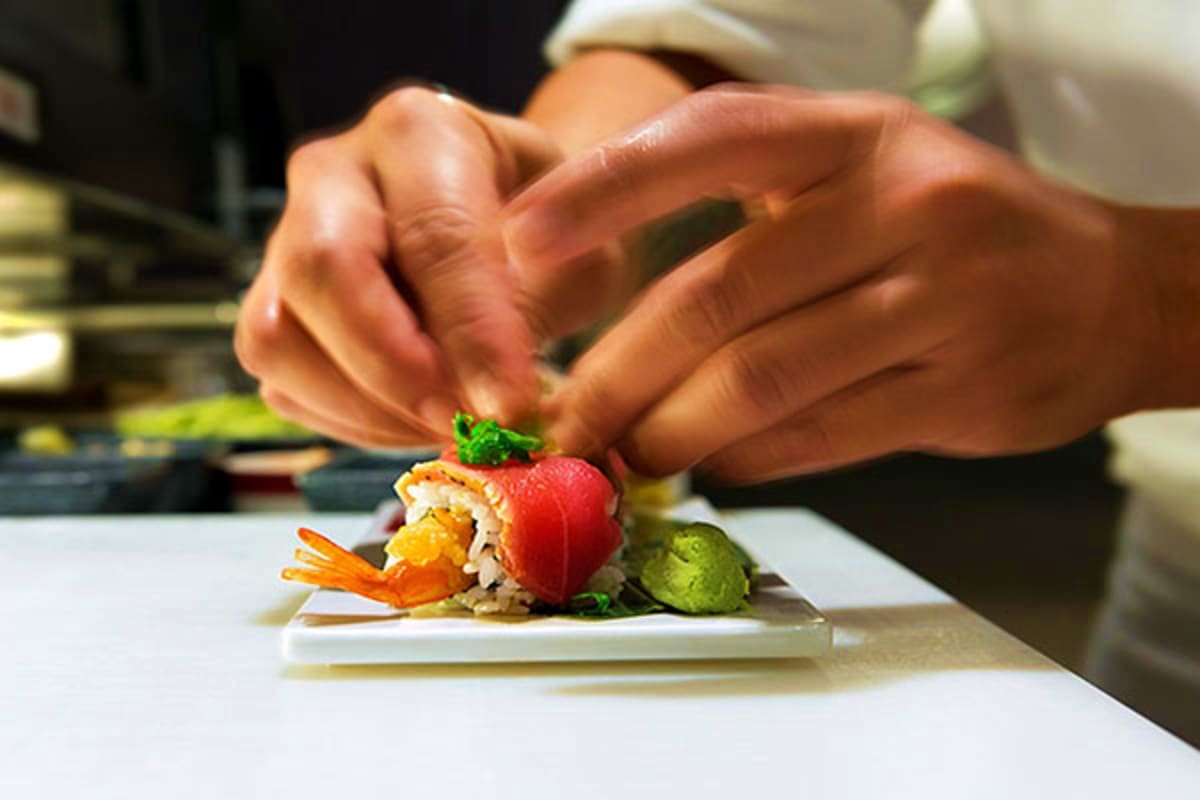 Chef's hands garnishing a fine dining sushi dish with microgreens and edible flowers on a white plate