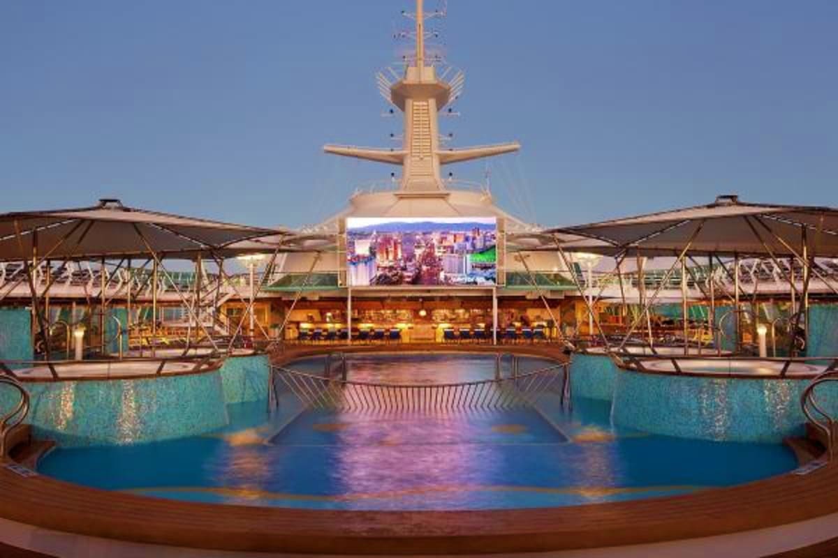 Cruise ship deck with tall central tower, blue pools, lounge seating areas under umbrellas, and digital display screen