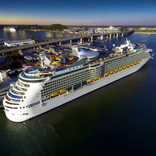 Aerial view of a large white cruise ship docked at a port terminal with blue water and coastal town buildings in background