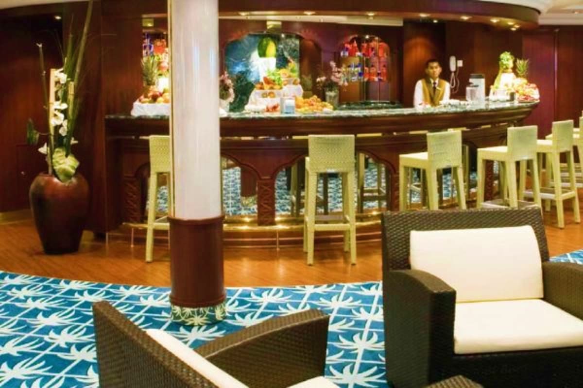Elegant cruise ship lounge with curved bar, cream bar stools, tropical plants, and decorative patterned flooring