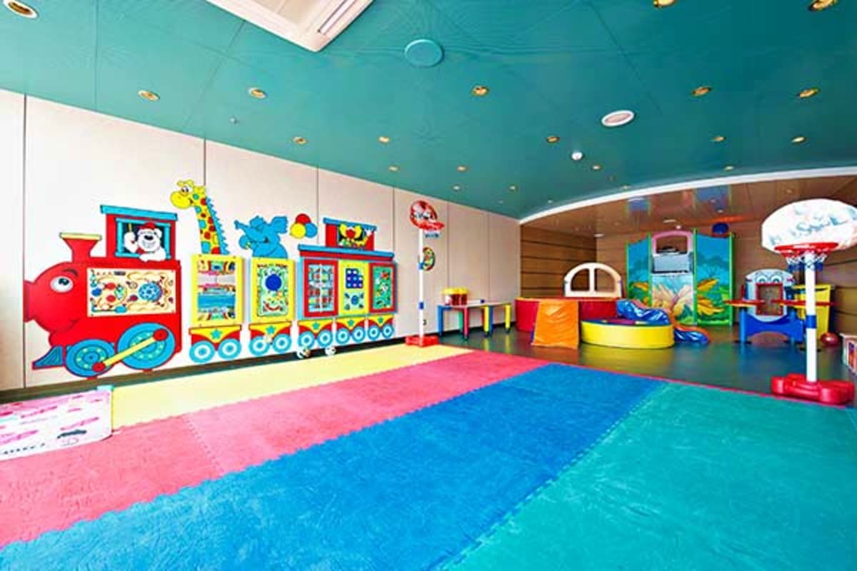 Colorful children's indoor play area with padded flooring, toy train wall mural, basketball hoop, and activity stations