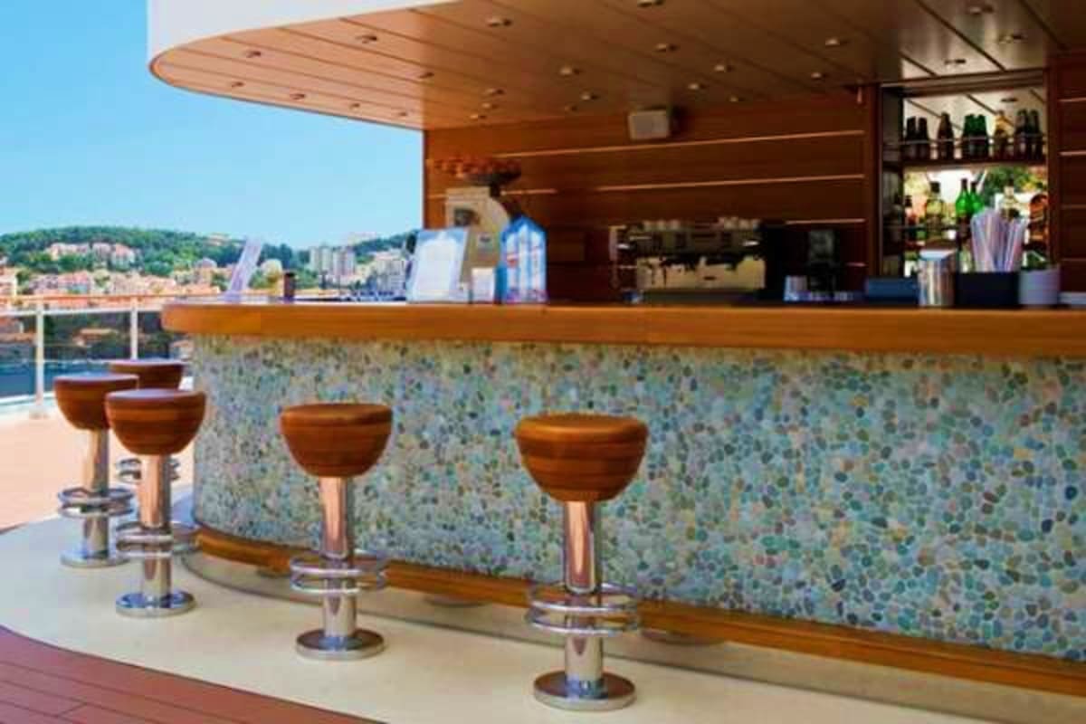 Modern cruise ship bar with wooden bowl-shaped stools, seafoam terrazzo counter, and Mediterranean coastal town visible throu