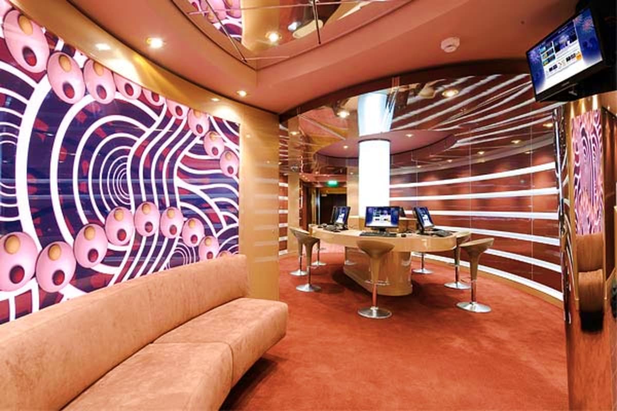Modern cruise ship interior with curved seating, illuminated geometric wall art, computer workstations, and contemporary loun
