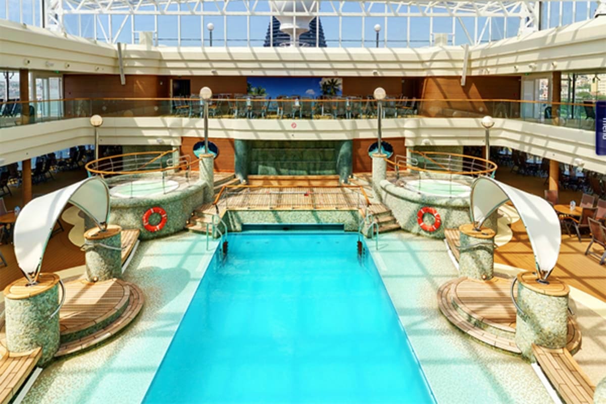 Luxury cruise ship indoor swimming pool with turquoise water, multi-deck atrium, curved seating areas, and glass ceiling on M